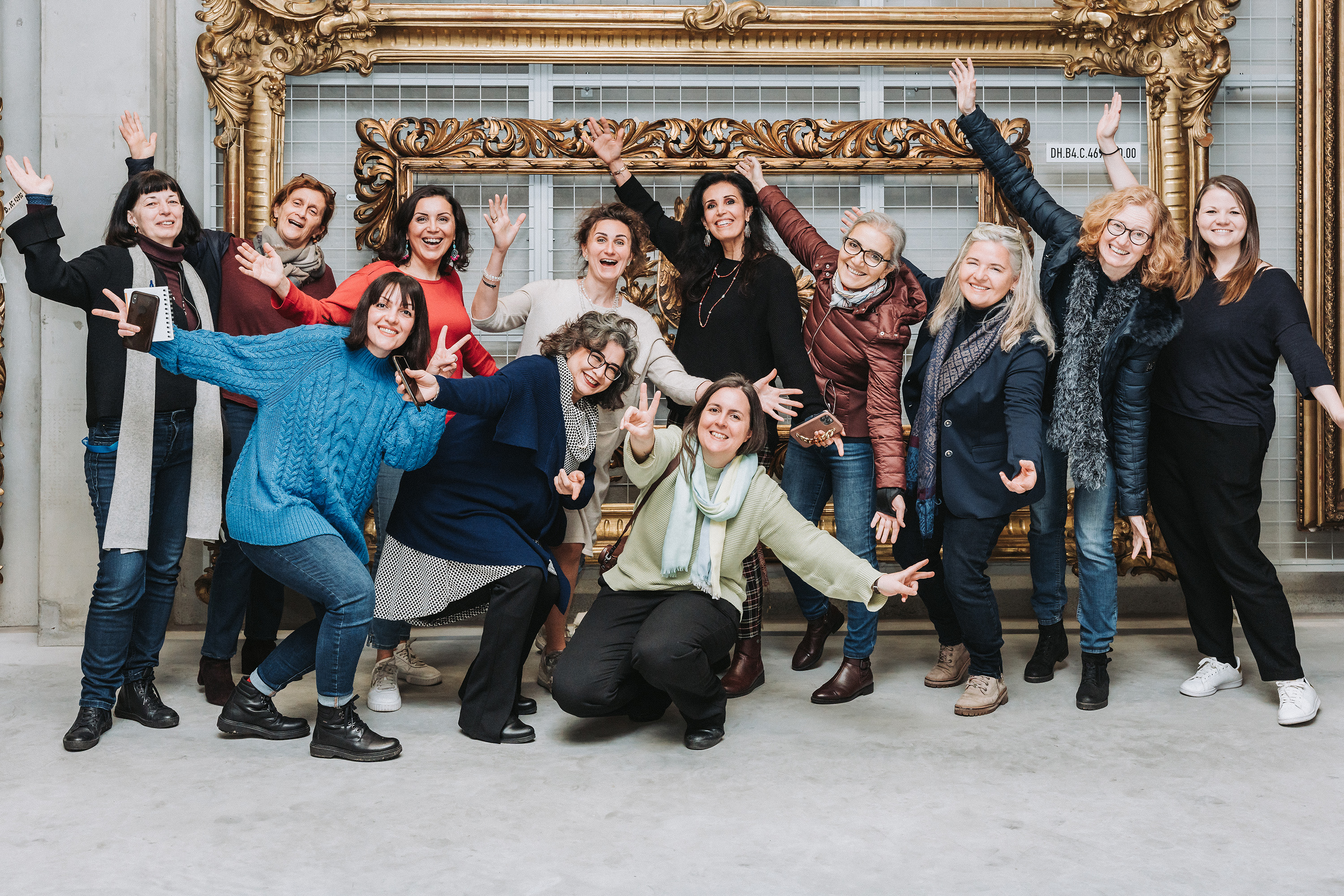 Gruppenfoto von 12 Kurator:innen die ihre Arme in die Luft strecken vor zwei goldenen Bilderrahmen.