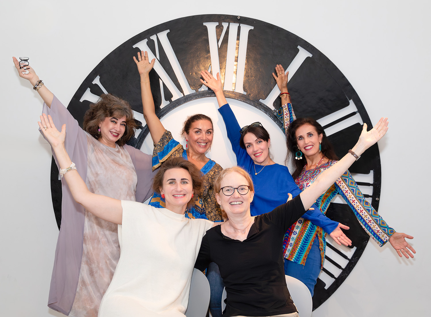 Gruppenfoto von 12 Kurator:innen die ihre Arme in die Luft strecken vor einem großen Ziffernblatt einer Uhr