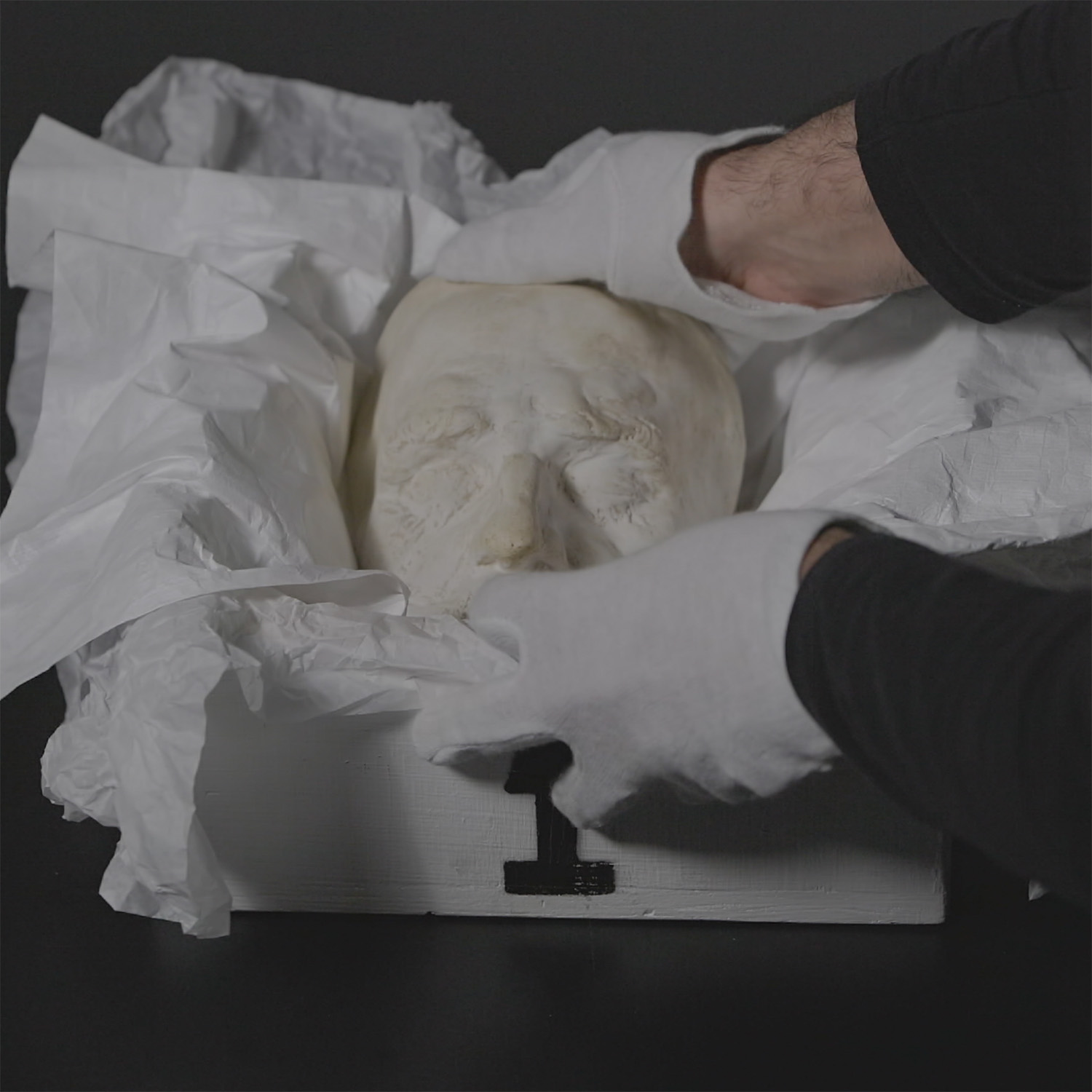 Totenmaske wird von zwei Händen mit weißen handschuhen in eine Kiste gelegt