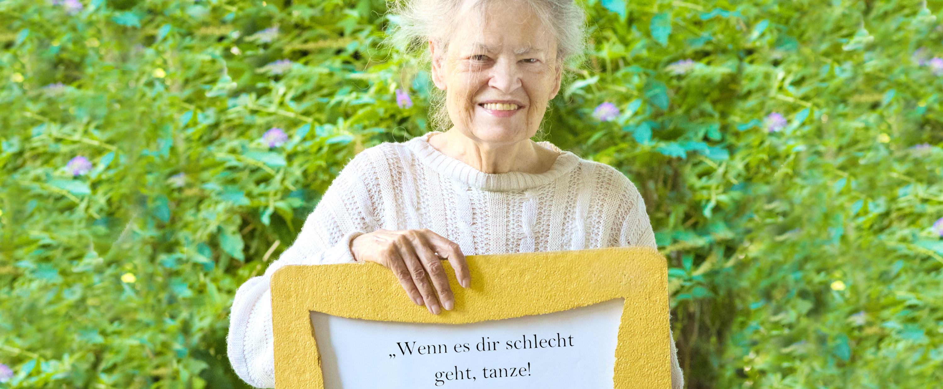 Ältere Frau hält ein gelbes Schild in der Hand mit dem Spruch: Mein Rat fürs Leben. Wenn es dir schlecht geht, tanze! Dann geht es dir wieder besser.