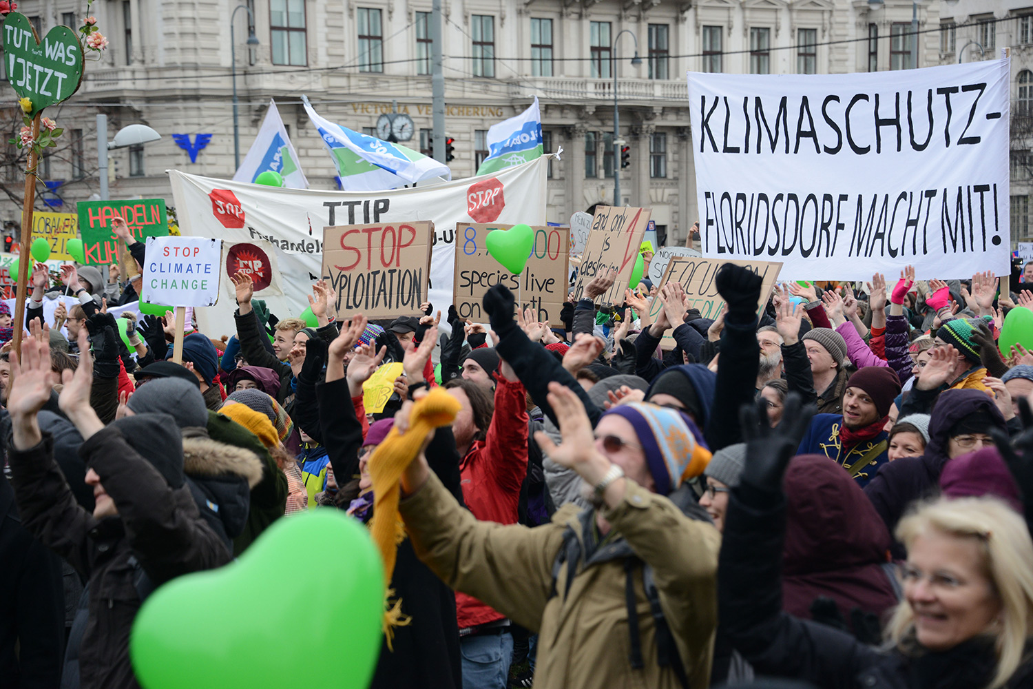 Eine große Menschenmenge bei einer Klimademonstration, die Schilder mit Aufschriften hochhält, zum Beispiel „Klimaschutz - Floridsdorf macht mit!“. Die Teilnehmenden tragen Winterkleidung, einige halten grüne Luftballons in Herzform.