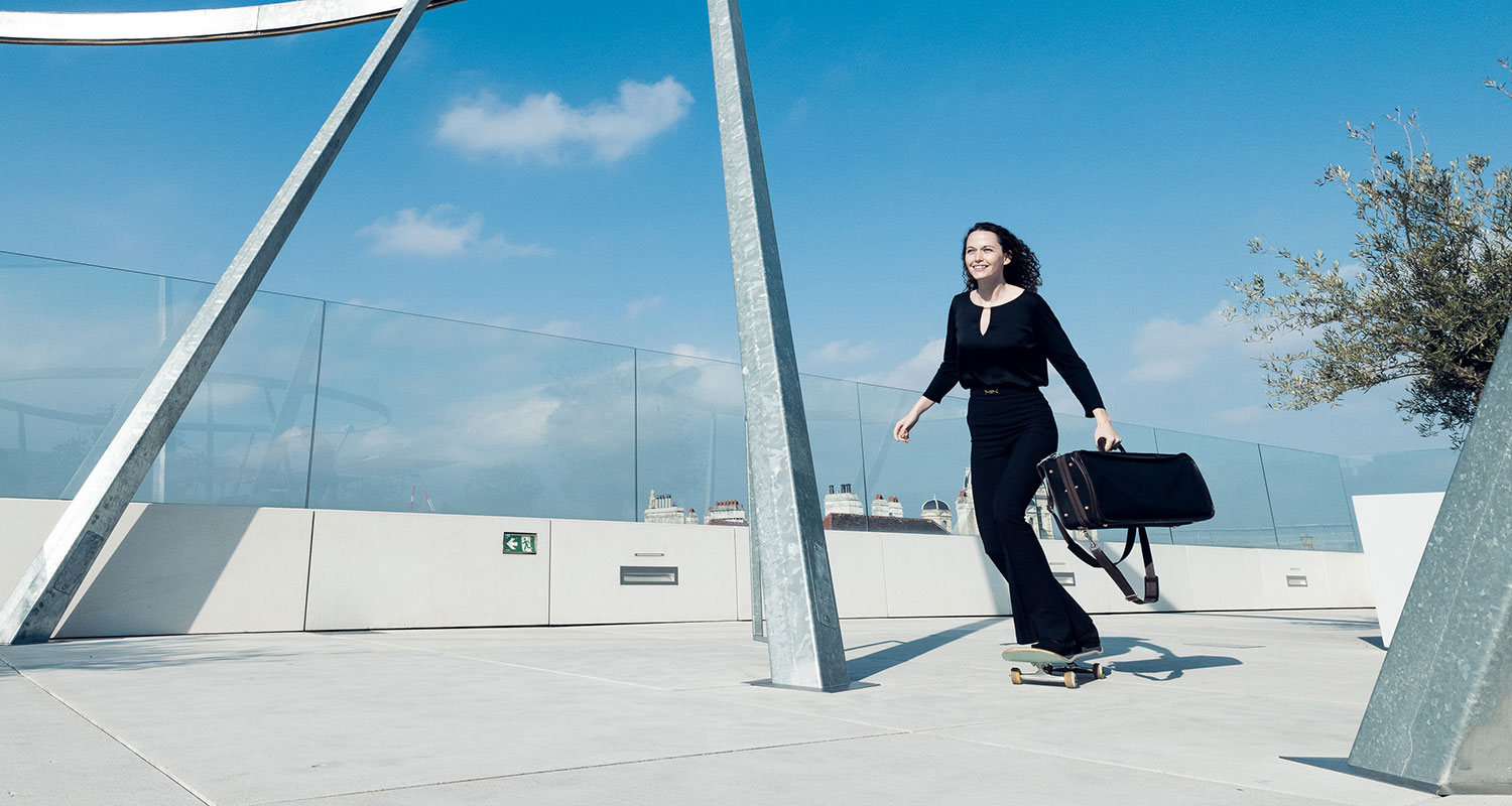 Musikerin mit Instrumentenkoffer auf einem Skateboard auf einer Dachterrasse