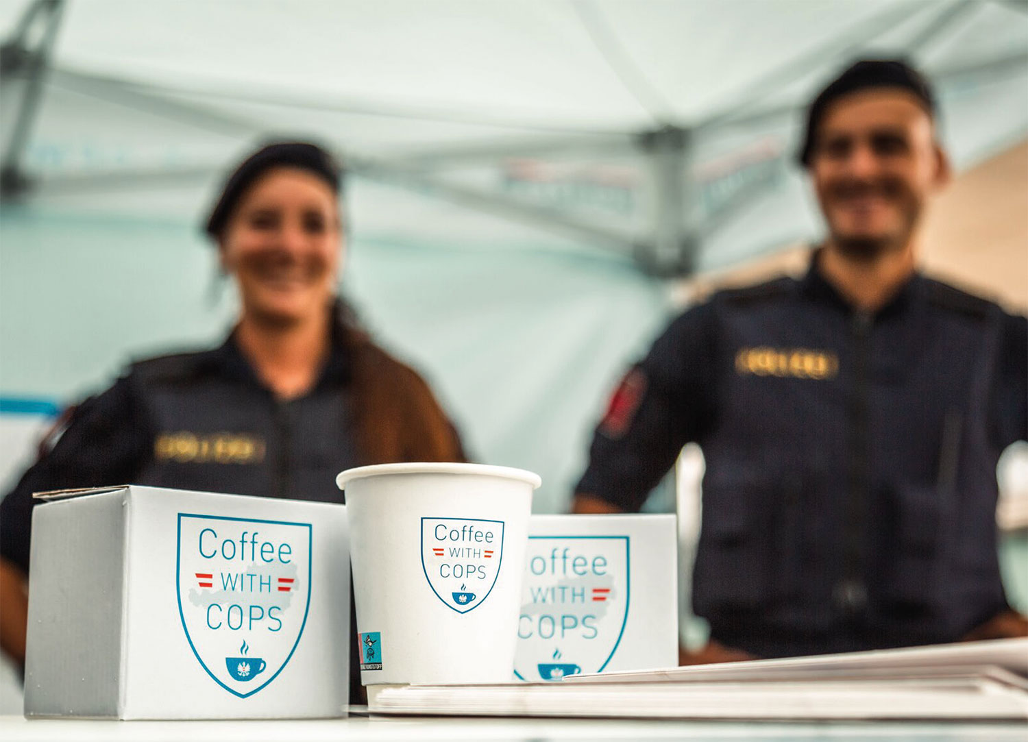 Tassen mit Aufdruck im Vordergrund dahinter eine Polizistin und ein Polizist in Uniform