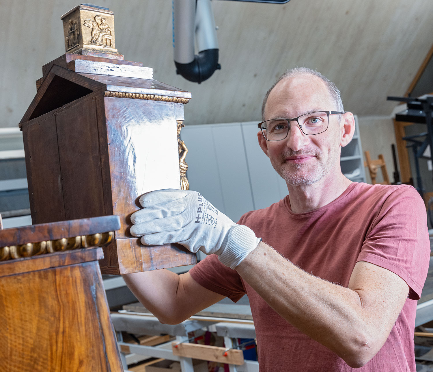 Der Restaurator steht in der Werkstatt und trägt mit Handschuhen einen Aufsatz eines Holzschrankes