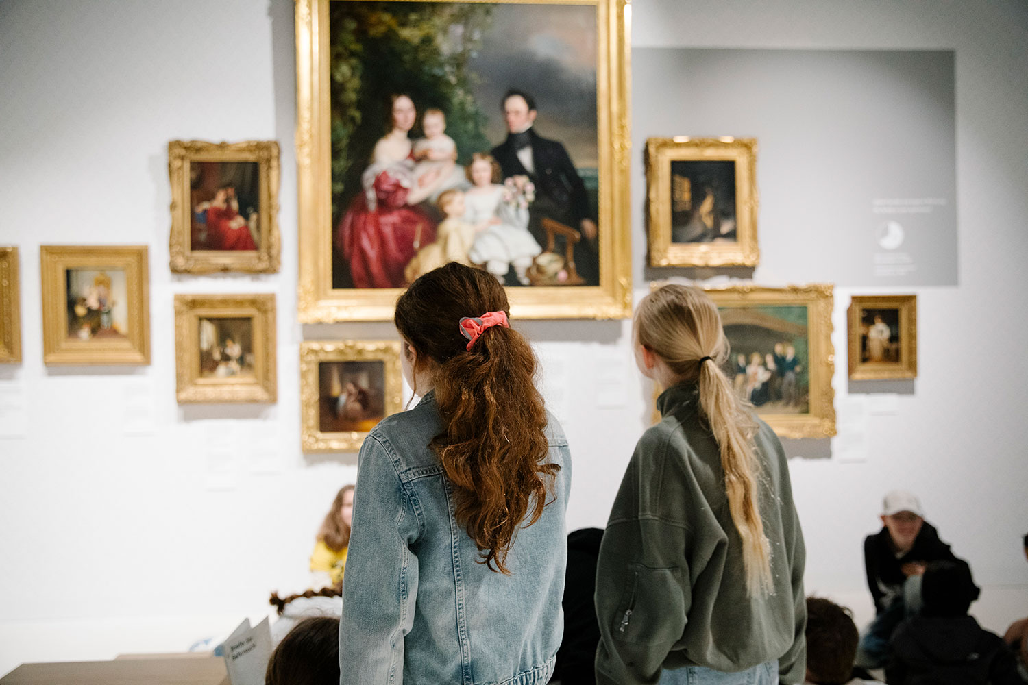 Ein blondes und ein braunhaariges Mädchen aus einer Schulgruppe stehen vor Waldmüller Gemälden 