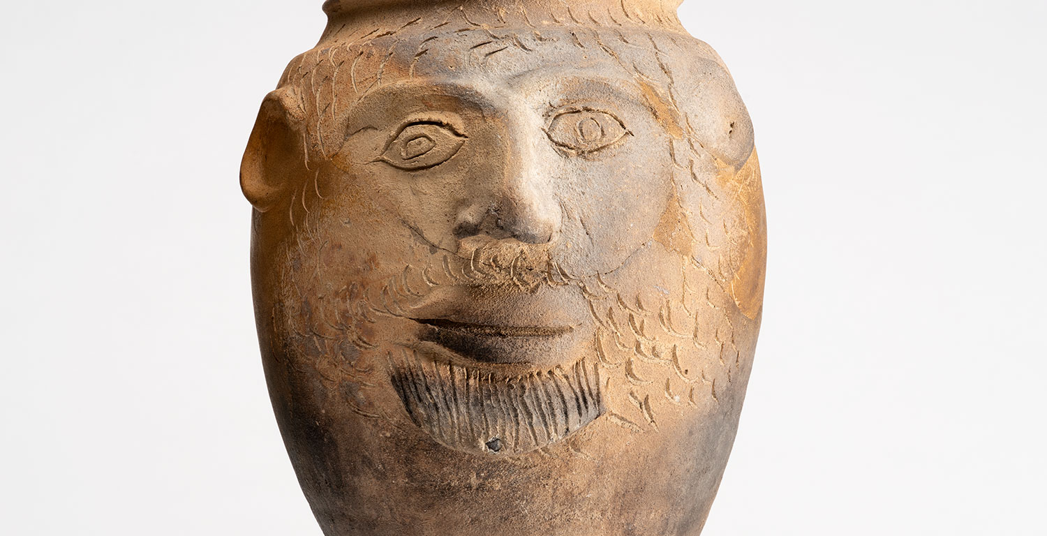 Eine braune Vase mit einem Gesicht und Ritzungen für Haare und Bart. Links ein gut sichtbares Ohr