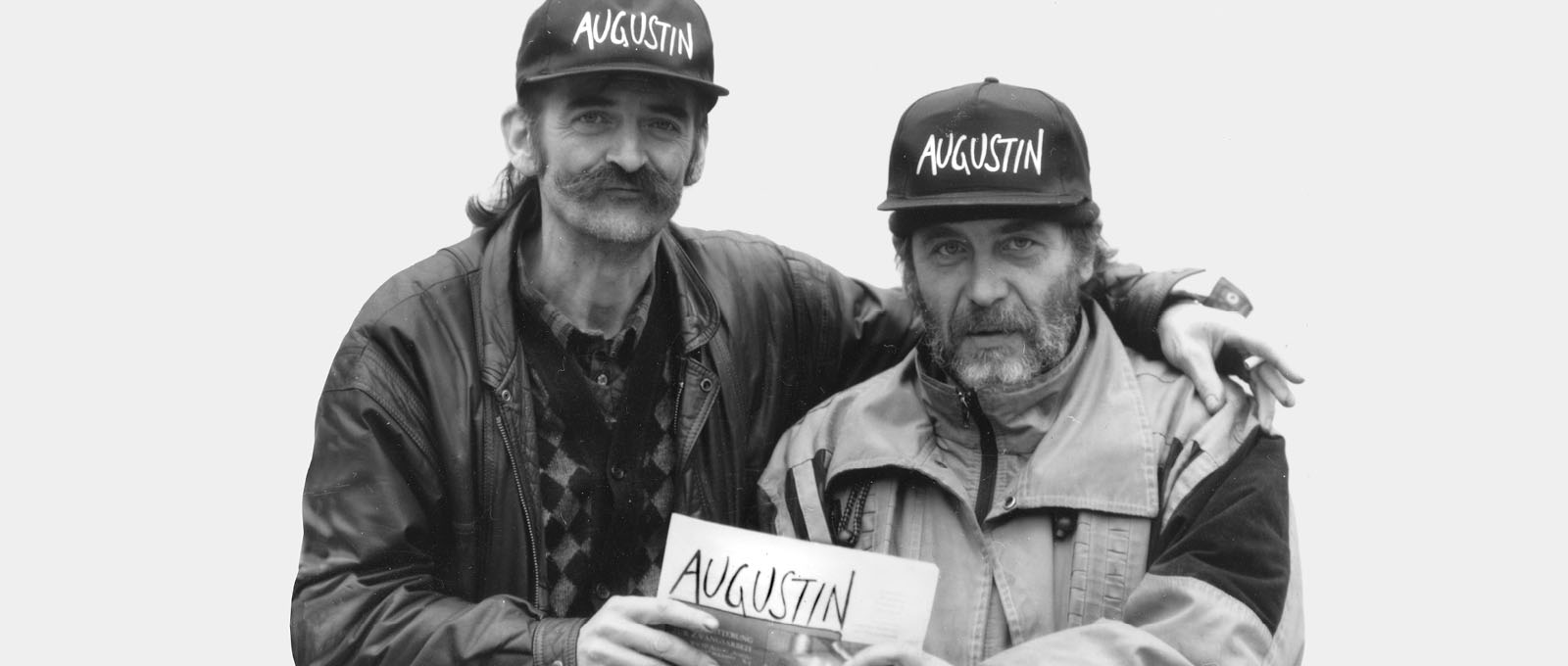 Zwei Augustinverkaufer in die Kameraschauend umarmen sich und halten eine Ausgabe des Augustin in der Hand.