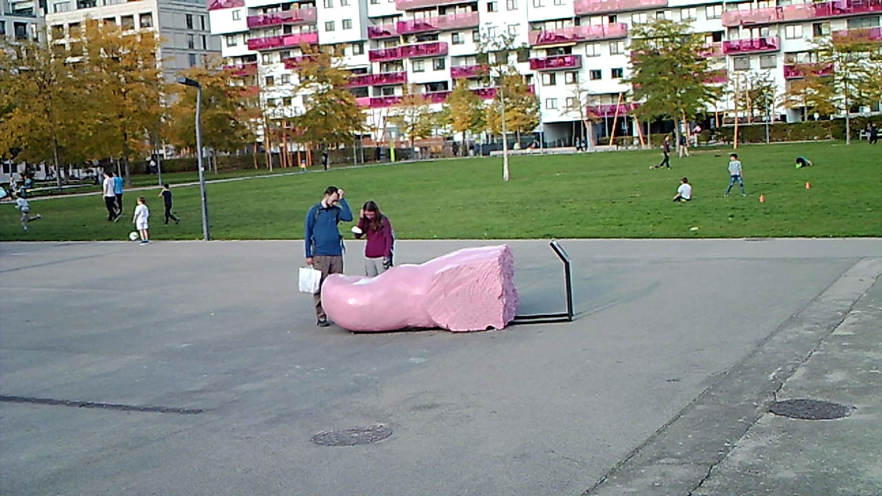 Großer rosa Zeh liegt auf einem Platz neben einem Fußballfeld auf dem Kinder spielen, ein Mann und eine Frau sehen den Zeh ratlos an