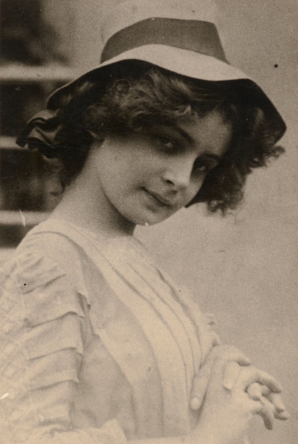 Junge Dame in einem hellen Kleid mit Falten in der Brustpartie, kurzen Haaren und einem Hut