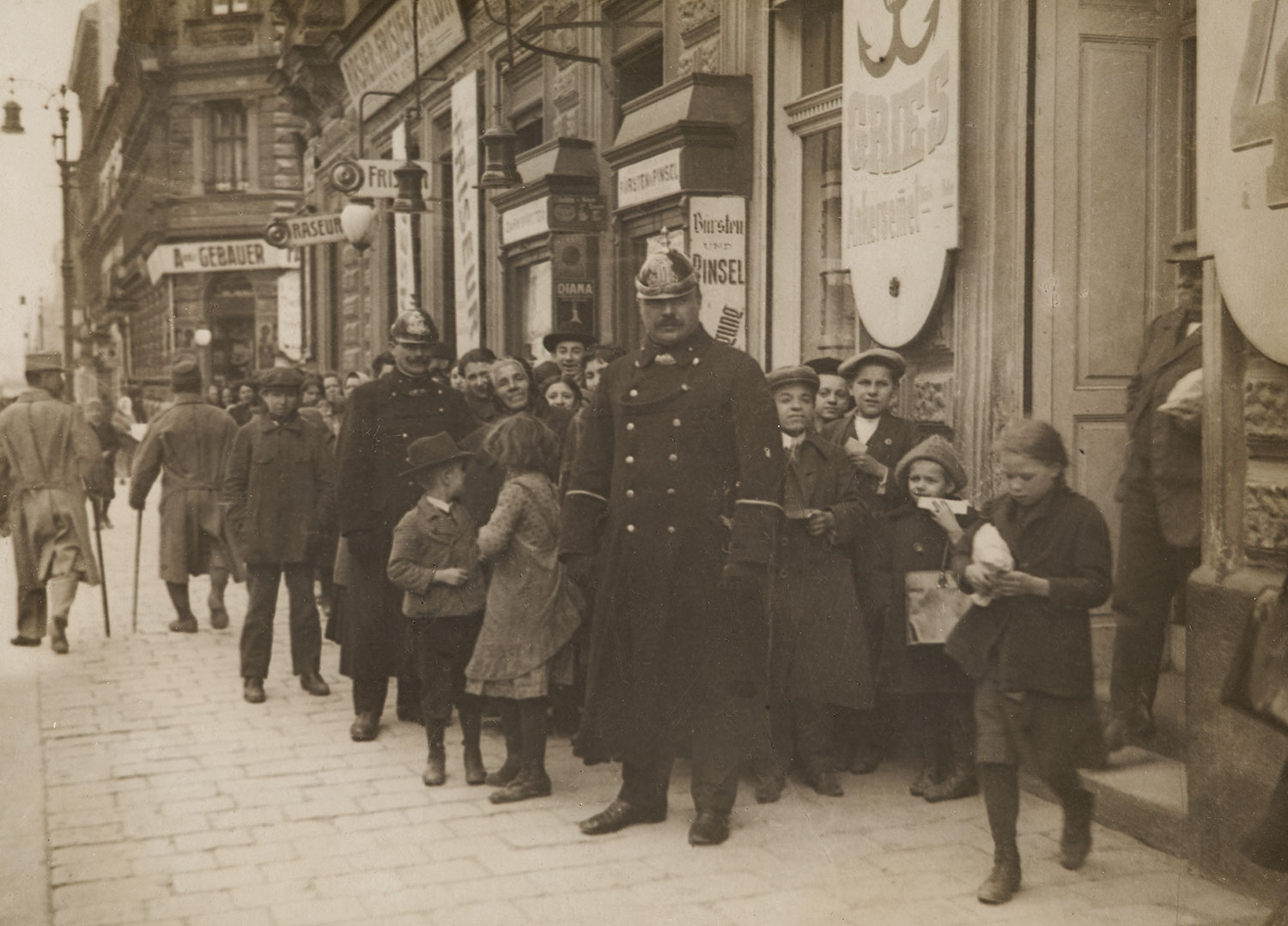Vor einem Geschäft steht ein Polizist in Uniform und Helm, umgeben von einer Menschenmenge, bestehend aus Erwachsenen und vielen Kindern.