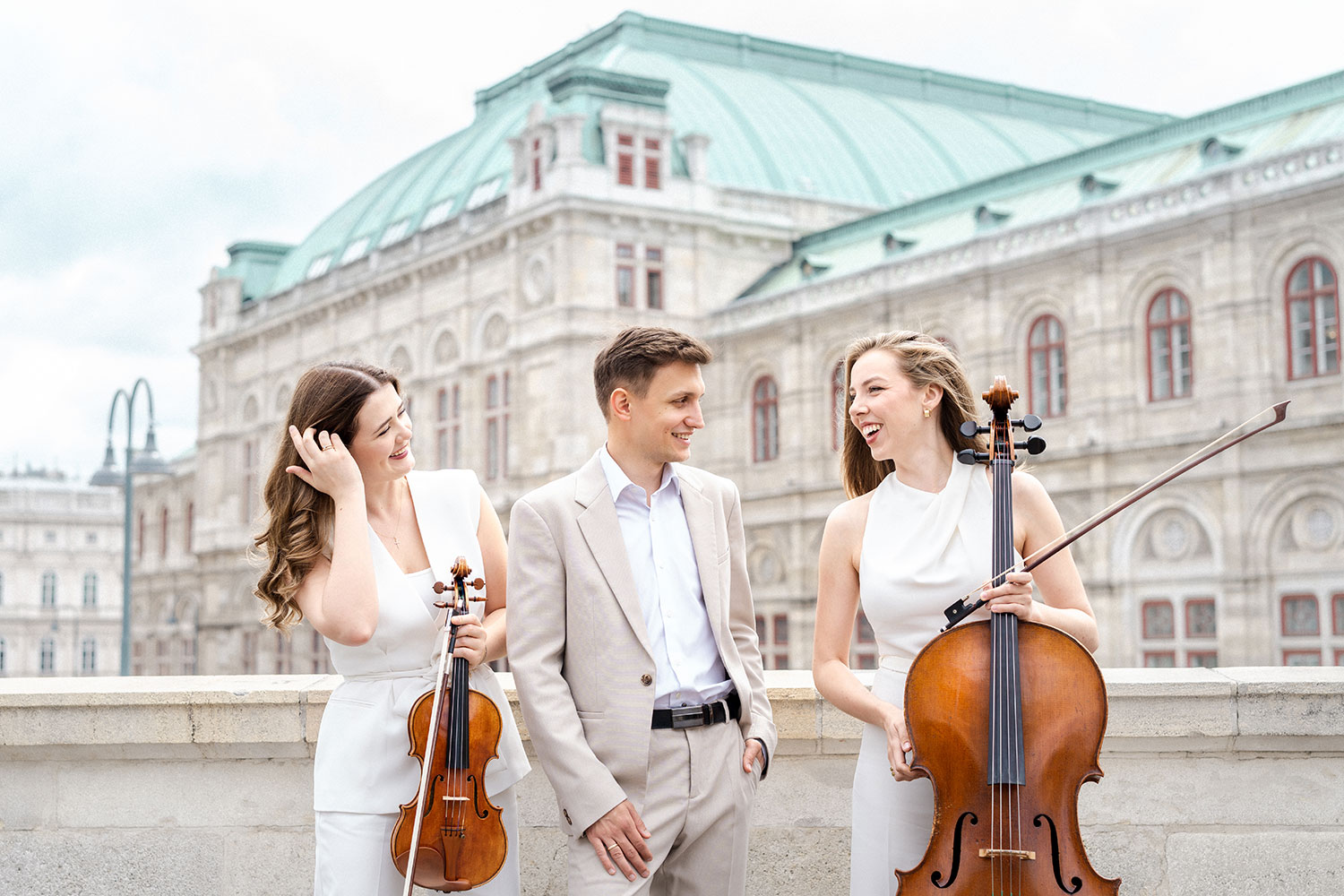 Zwei Musikerinnen mit ihren Instrumenten und ein Mann stehen erhöht mit der Wiener Staatsoper im Hintergrund