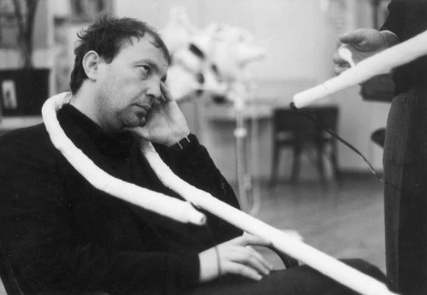 Franz West sitzt auf einem Stuhl und blickt nach oben, um seinen Hals ist ein skulpturales weißes Rohr geschlungen 