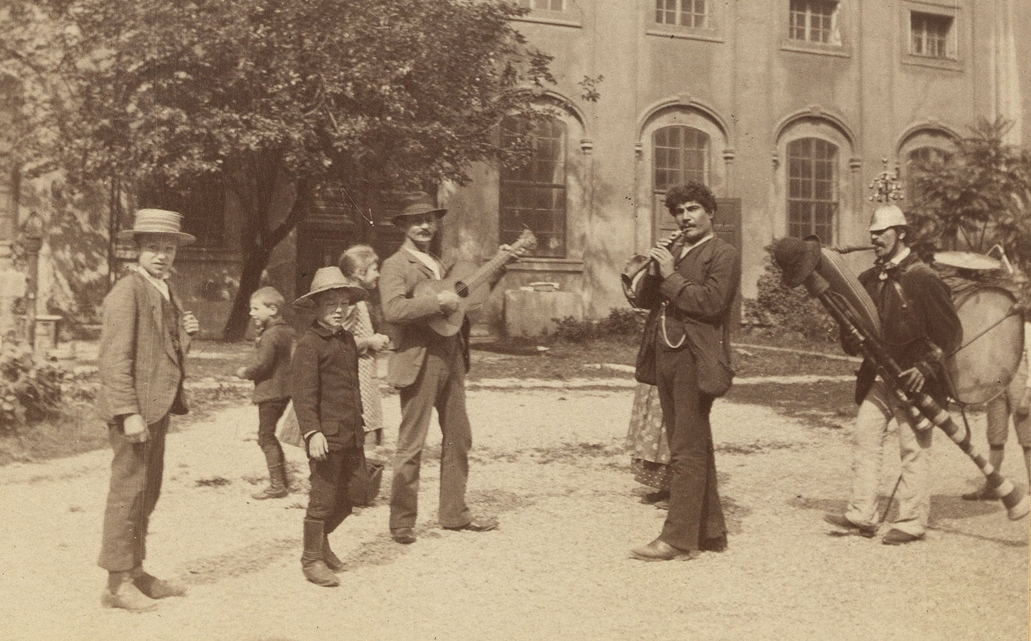 Drei Männer mit Instrumenten und Kinder unter einem Baum und vor einem Gebäude