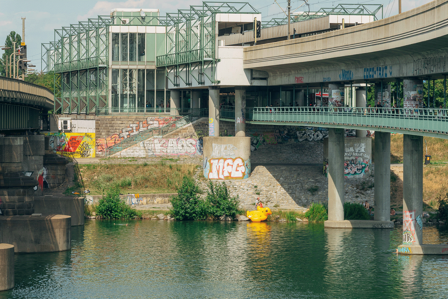 Steg über die Donau, Mauern mit Graffiti und eine Person am Ufer mit einer großen aufbblasbaren gelben Ente