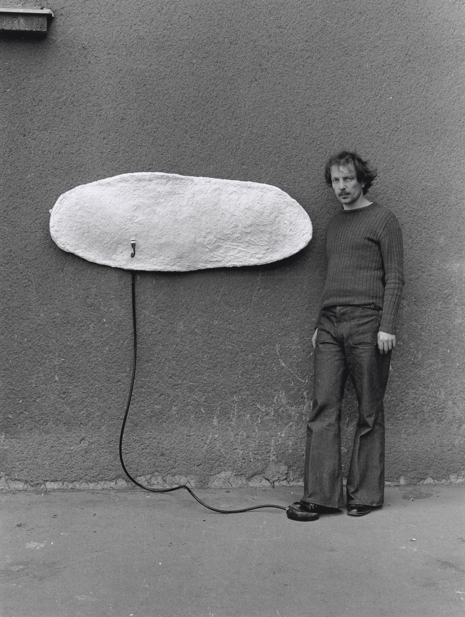 Franz West lehnt an einer Wand an der  sein Kunstwerk hängt